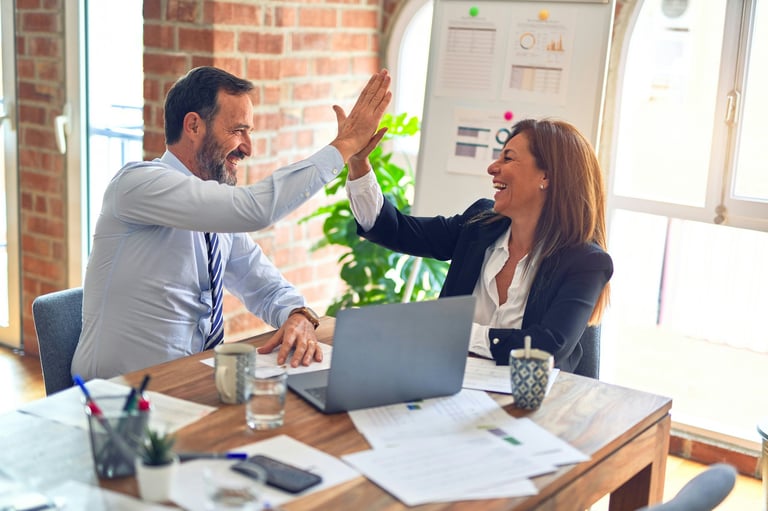 Two professionals high-fiving at a desk with laptop and papers in a bright office with brick walls