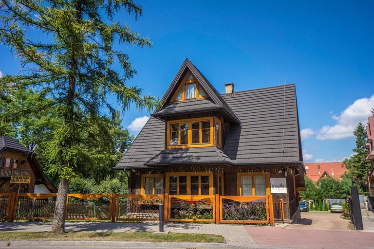 Charming two-story wooden house with dark roof, yellow windows, and wooden fence on a sunny day with blue sky and trees