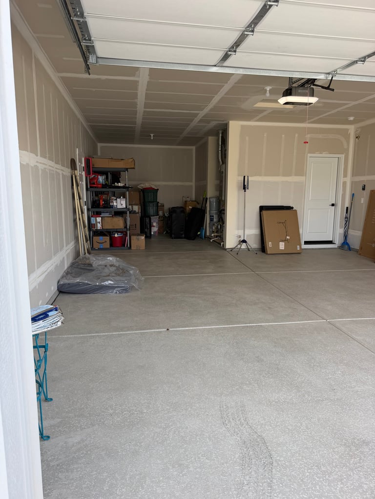 Interior of empty garage with concrete floor, white walls, overhead door, and storage shelves along the sides.