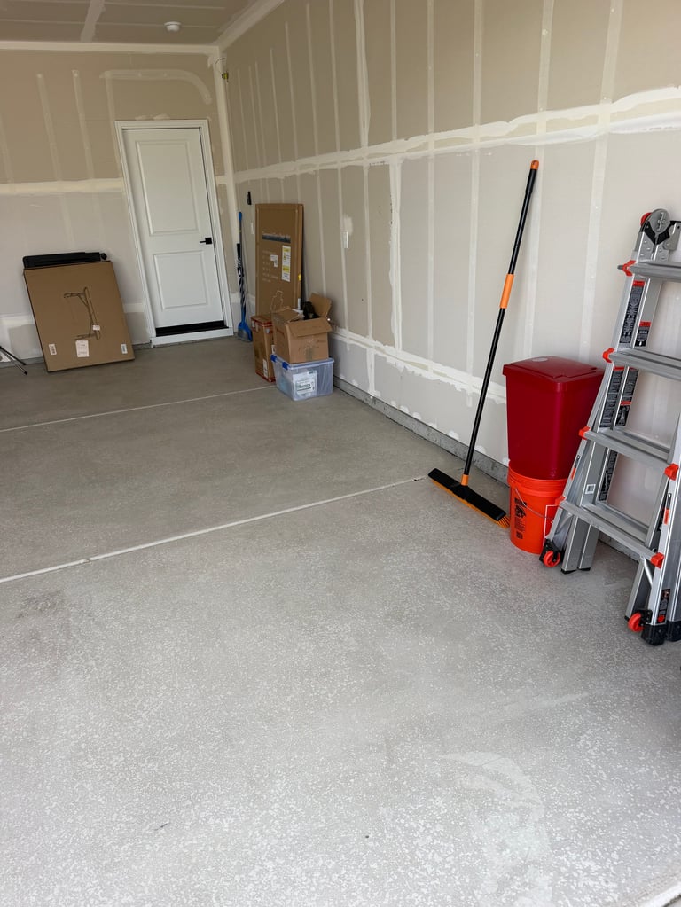 Unfinished garage interior with white drywall, concrete floor, cardboard boxes, red storage containers, ladder, and broom during construction.