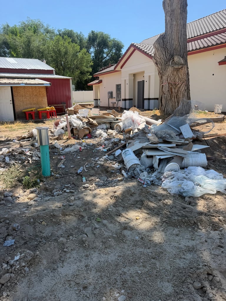 Construction site with scattered debris, PVC pipes, and building materials in front of residential homes with trees.