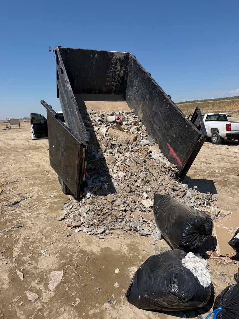 Dump truck unloading construction debris and rubble onto a dirt lot with black garbage bags nearby.