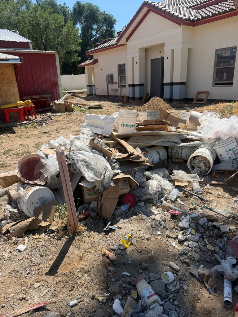 Construction debris and waste materials scattered on bare ground in front of a modern house under renovation.