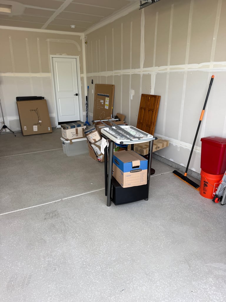 Unfinished garage interior with beige and white paneled walls, concrete floor, and various cardboard boxes, wooden panels, and red storage items scattered throughout.
