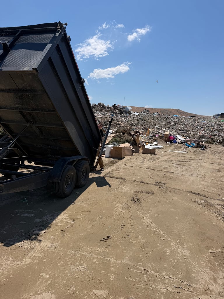 A dump truck with its bed raised unloads debris at a landfill site with piles of waste under a blue sky.