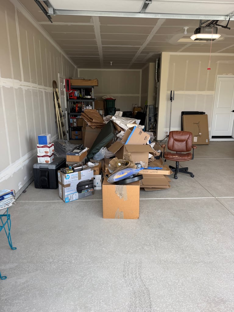 Cluttered garage interior with cardboard boxes, plastic bins, a red leather chair, and various items scattered on the concrete floor.