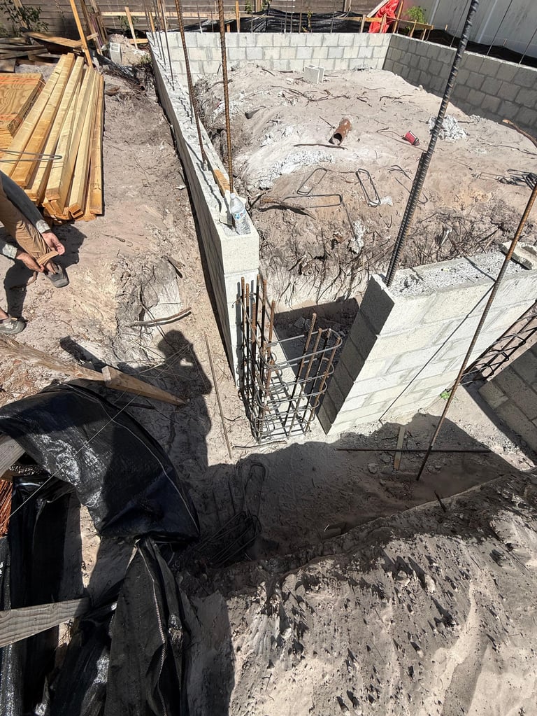 Construction site with open excavation pit, metal reinforcement bars, lumber, and two workers in sandy terrain