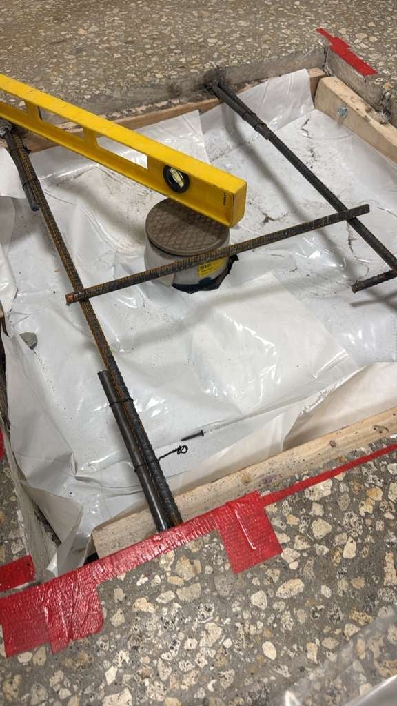 Construction tools in a white bucket including a yellow level, rebar, and a round tool, positioned on concrete