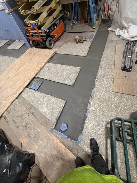 Warehouse or workshop floor showing flooring installation with light wood planks and gray underlayment, construction equipment and tools visible