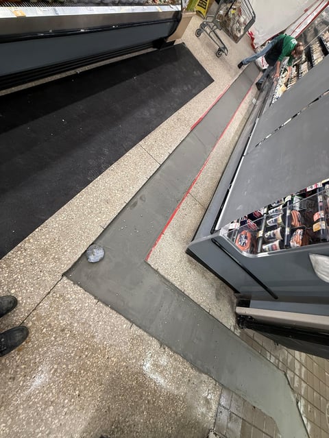 Overhead view of a storefront entrance with black awning, red striped line, and postcard display stand on the sidewalk