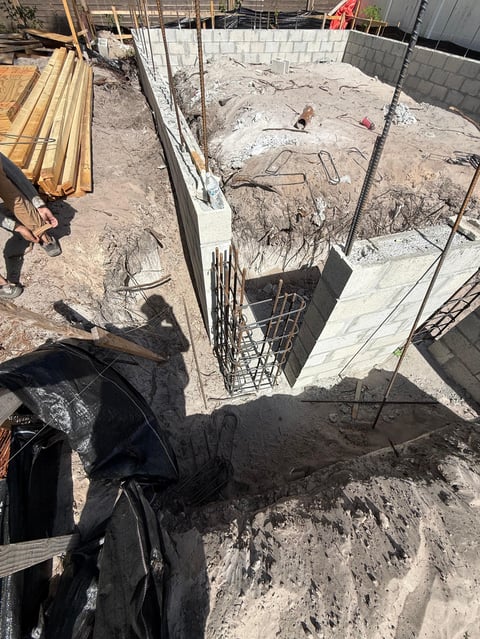 Construction site with excavated basement pit, metal rebar cage, lumber stacks, and workers preparing concrete foundation for building