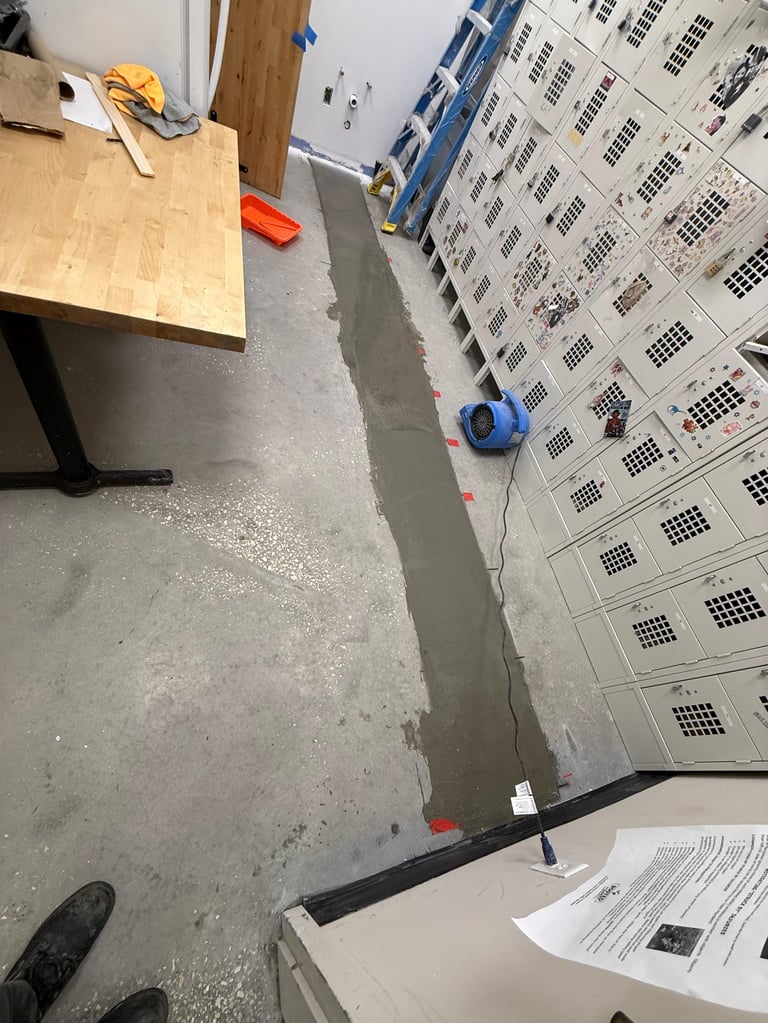 Overhead view of concrete floor with wet epoxy coating being applied, showing a wooden workbench, plastic storage boxes, and construction materials