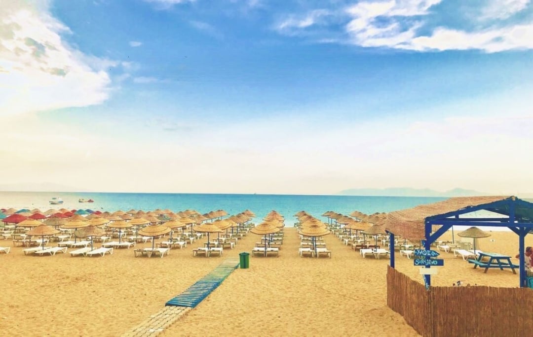 Organized beach with rows of beach chairs and umbrellas, blue shelters, calm turquoise sea, and clear sky