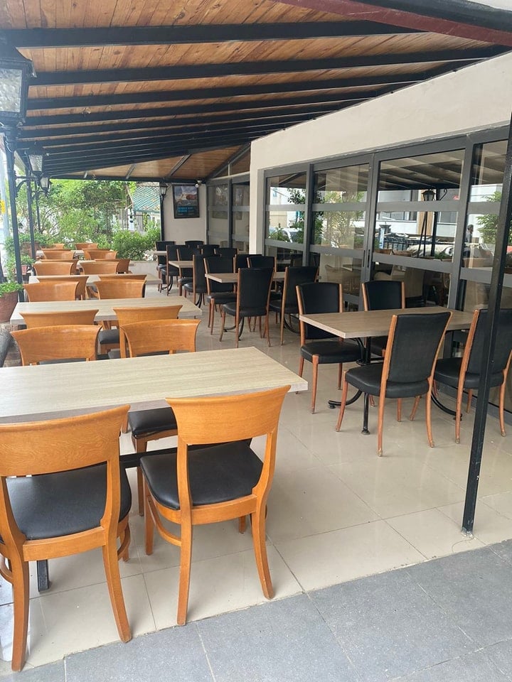 Modern restaurant patio with wooden beams, tables with wooden chairs and black cushioned seats, glass doors, and potted plants