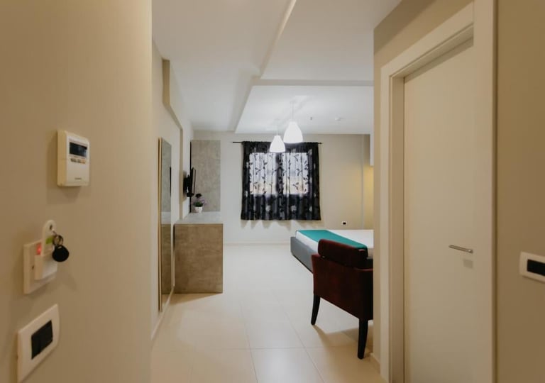 Modern hallway with white polished concrete floor, beige walls, wall-mounted controls, and furniture including a dark desk and turquoise chair