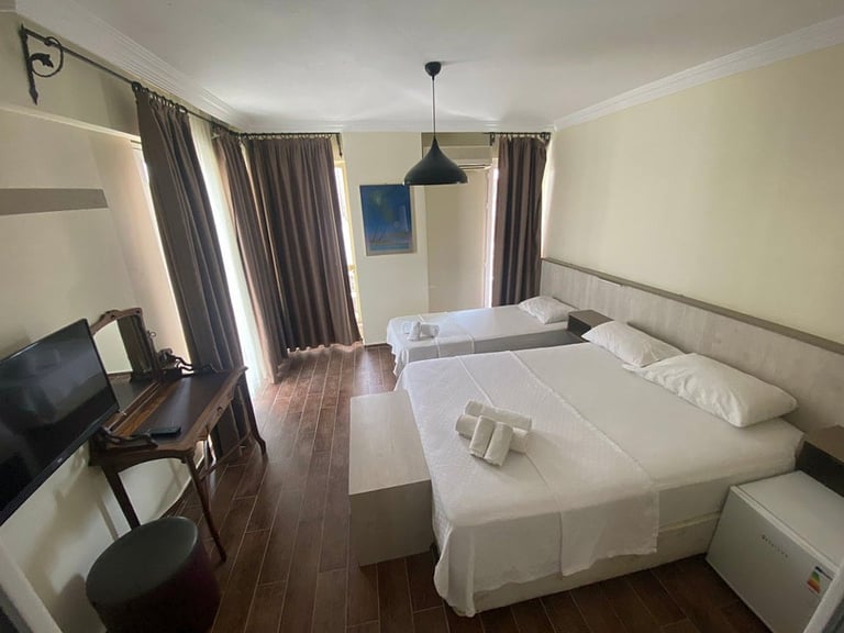 Hotel room interior with two twin beds, desk with TV, windows with curtains, and dark wood flooring