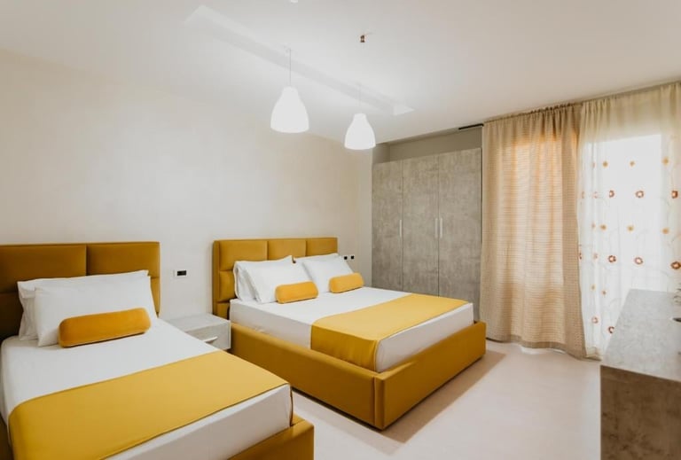 Modern bedroom with two mustard-yellow upholstered beds, white bedding, pendant lights, and beige curtains