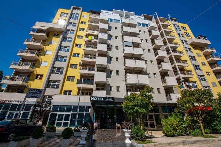 Modern multi-story hotel building with yellow and white striped facade, balconies, and ground-floor entrance against blue sky