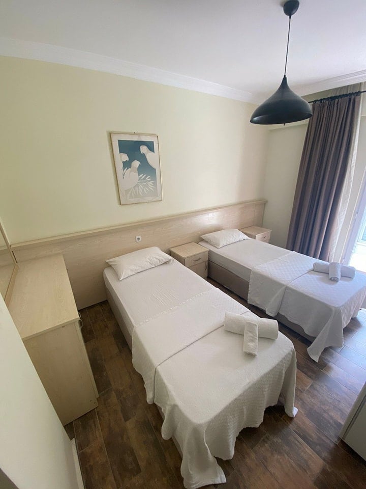 Hotel room with three twin beds with white linens, yellow walls, wood flooring, black pendant light, and framed artwork