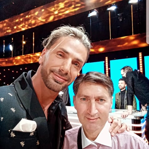 Two men posing together on a TV studio set with illuminated backdrop and other people visible in background