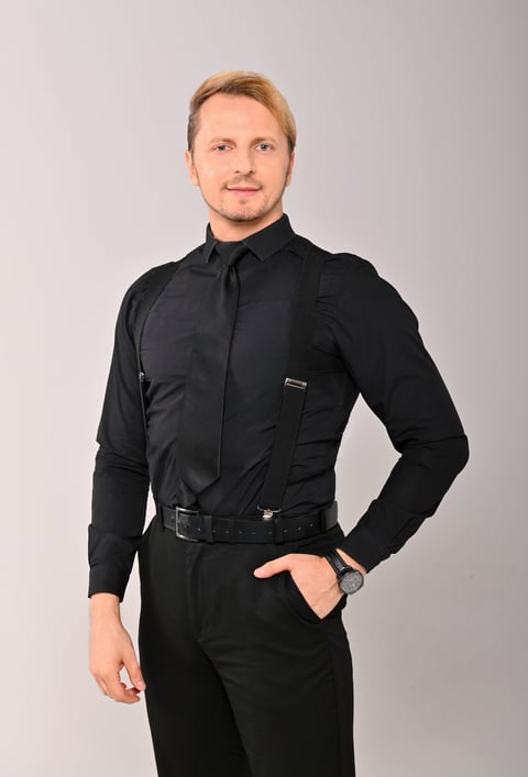 Professional headshot of man wearing black shirt, tie and pants with hand in pocket against neutral background