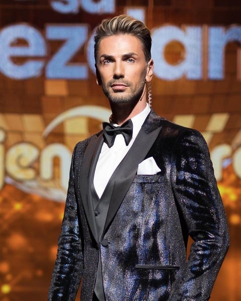 Man in navy sequined tuxedo with black bow tie at formal event, standing against illuminated backdrop
