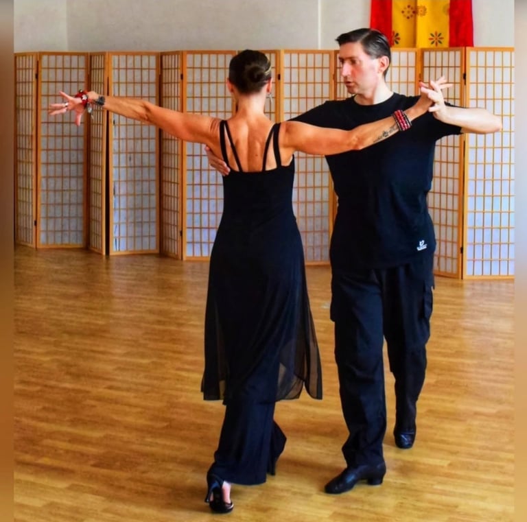 Two ballroom dancers in black attire performing a dance move with arms extended in a studio with wooden flooring