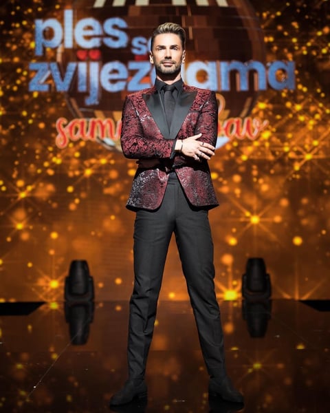 Man in burgundy sparkly blazer and black pants standing with crossed arms on a golden-lit stage