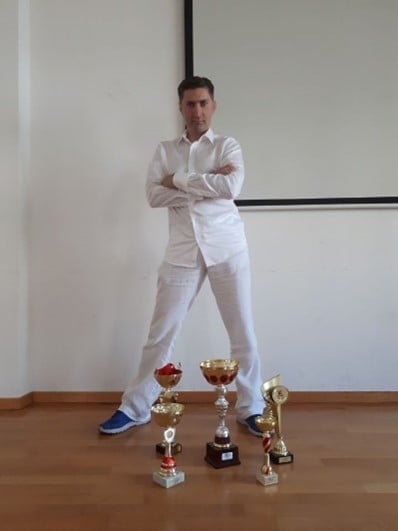 Man in white outfit standing with arms crossed in front of multiple trophy cups on wooden floor