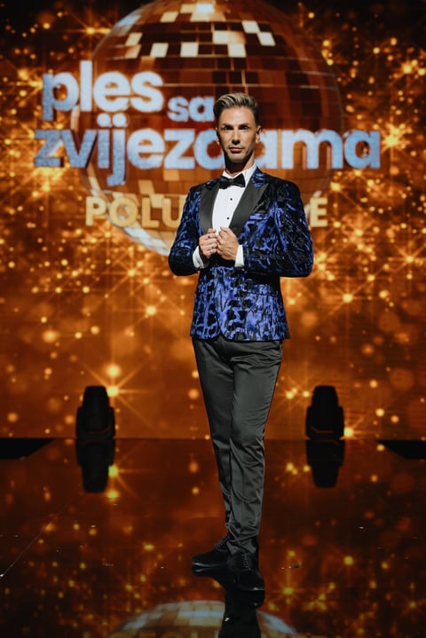 Man in blue sequined blazer and black pants standing on stage with golden disco ball and sparkles behind him