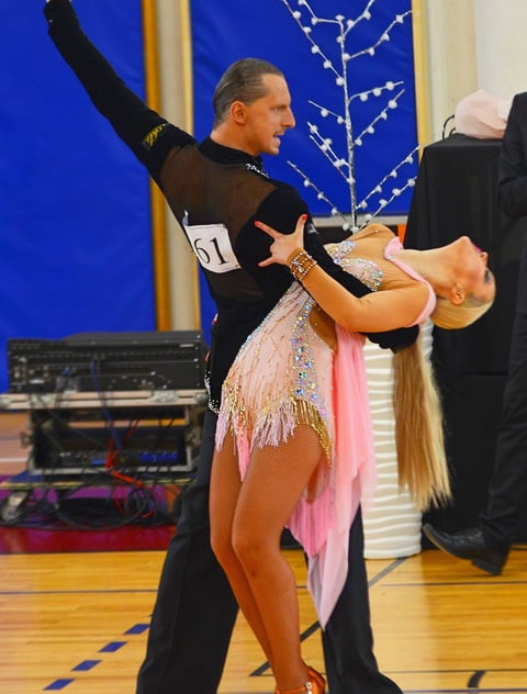 Male and female ballroom dancers performing a competition routine, with the man lifting the woman in a pink sequined dress on a wooden dance floor