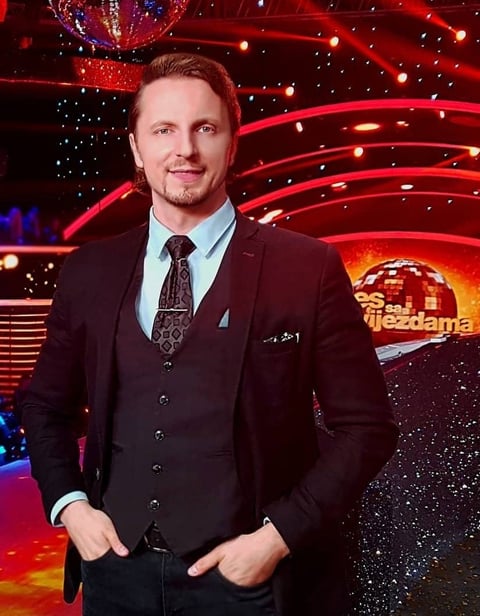 Man in dark suit and light blue shirt smiling at camera with red neon stage backdrop and disco ball behind him