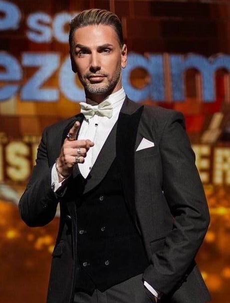 Man in formal black tuxedo with white bow tie holding microphone on stage with colorful backdrop
