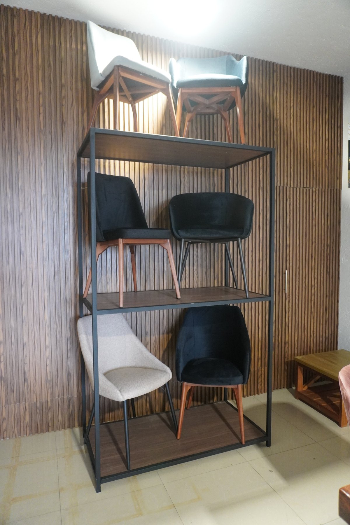 Industrial metal shelving unit displaying various modern chairs with black and wooden frames against corrugated wall backdrop