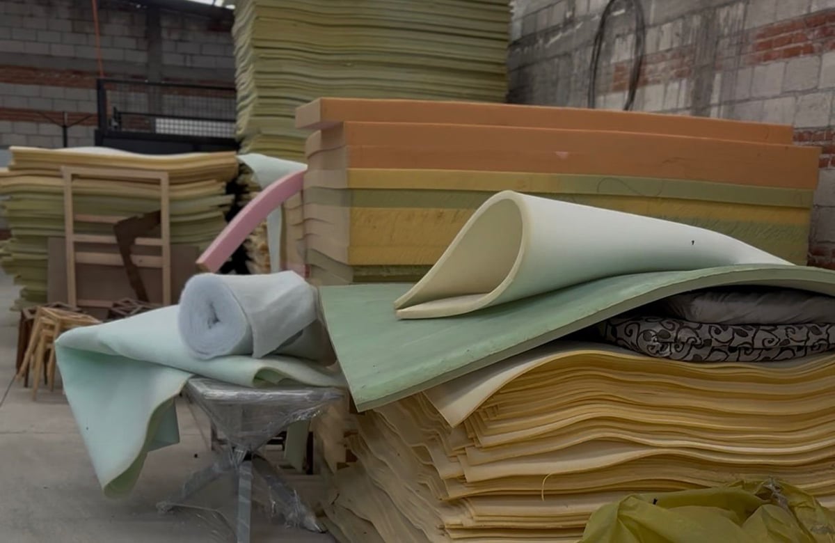 Warehouse storage area with stacked foam mattresses, rolled upholstery materials, and wood-framed furniture pieces in various pastel colors.