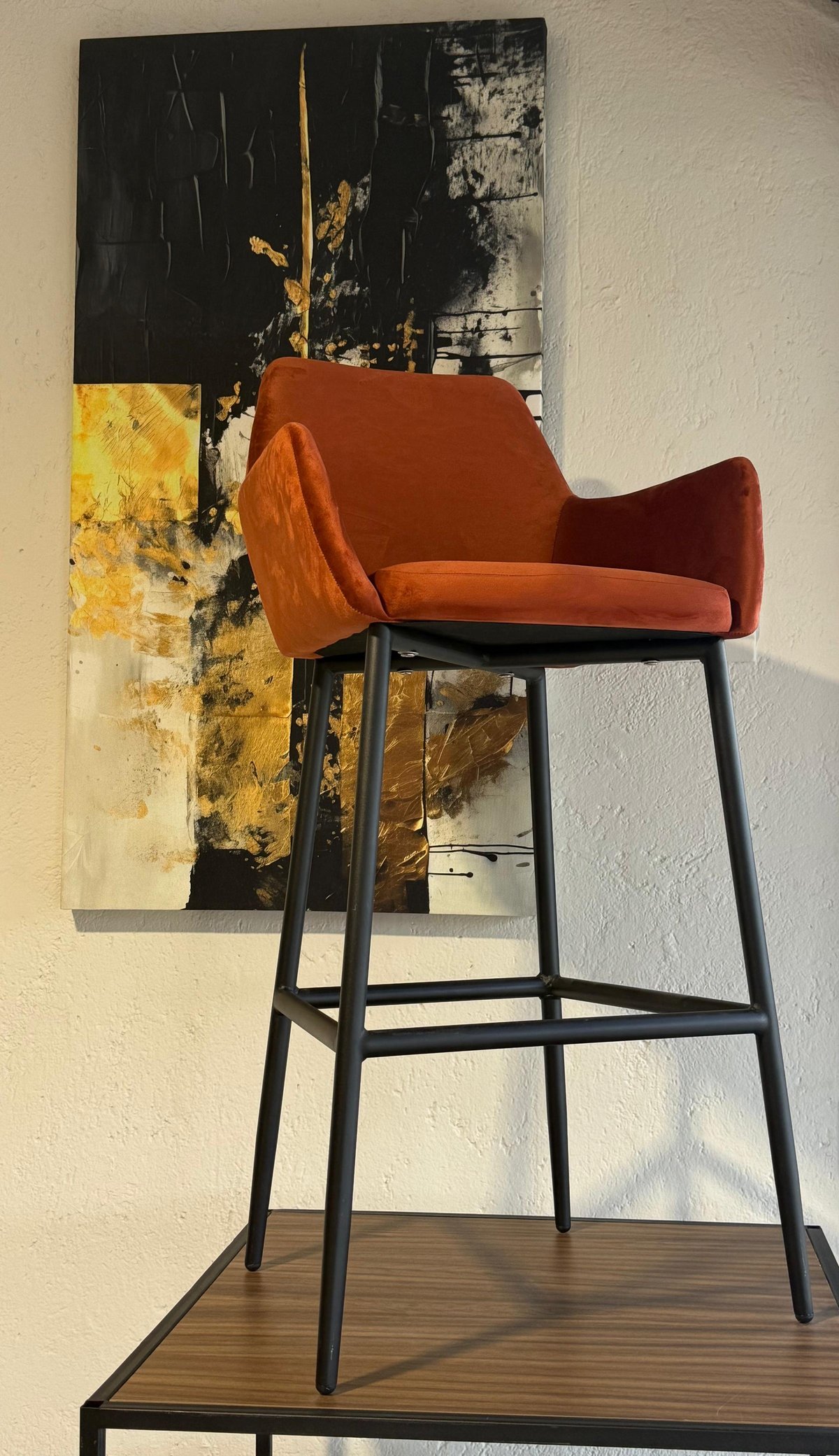 Orange modern bar stool with curved armrests and black metal legs, displayed against an abstract painting with black, yellow, and gold tones.