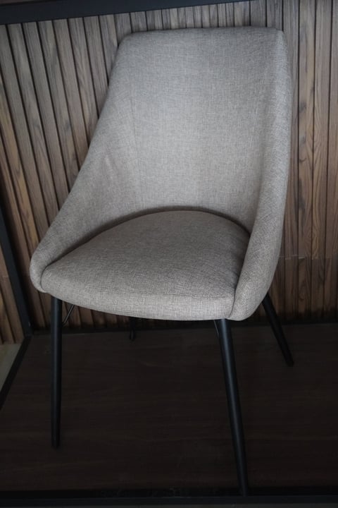 Modern upholstered dining chair with light gray fabric seat, curved backrest, and black metal legs against a wood-paneled wall