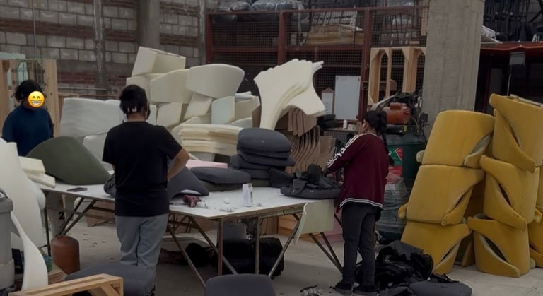 People working in a warehouse surrounded by stacked white furniture, yellow cushions, and cardboard boxes
