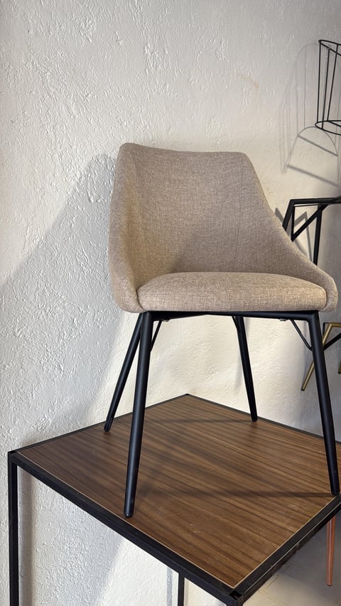 Beige upholstered chair with black metal frame displayed on dark wood table against white textured wall