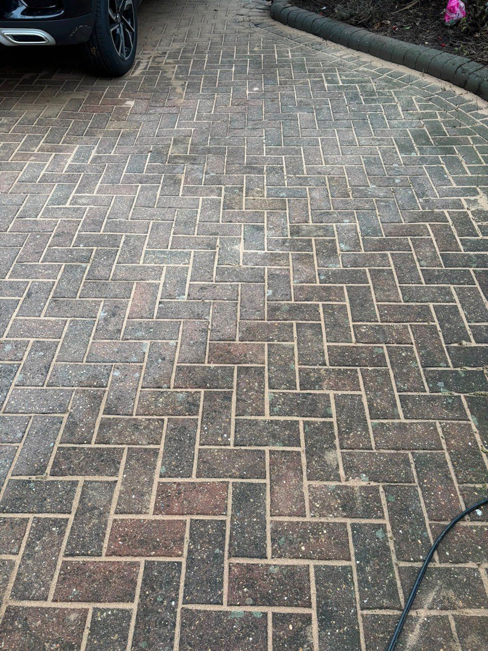 Herringbone patterned brick driveway with moss growth, bordered by a parked car and lawn edge