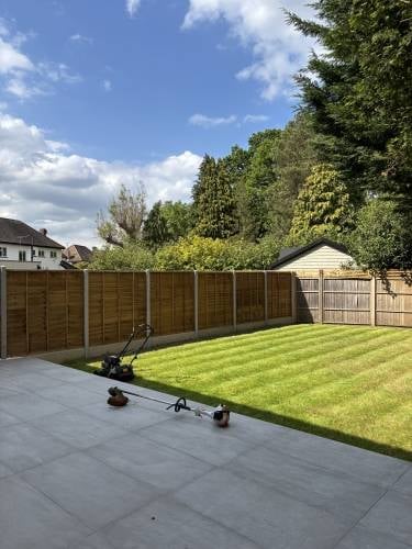 Modern backyard garden with patio, striped lawn, wooden fencing, and garden shed under blue sky