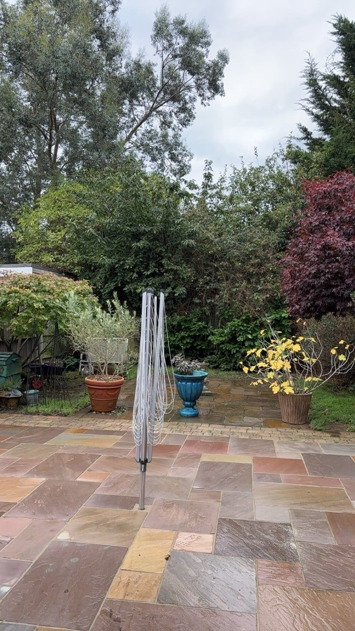 Closed umbrella with gray cover standing on multicolored stone patio in a garden with potted plants and trees