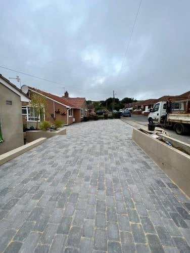 Newly paved residential driveway with gray pavers leading through a village street lined with brick homes and parked vehicles