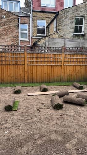 Backyard with wooden fence, scattered logs and wooden planks on bare ground, brick houses visible in background