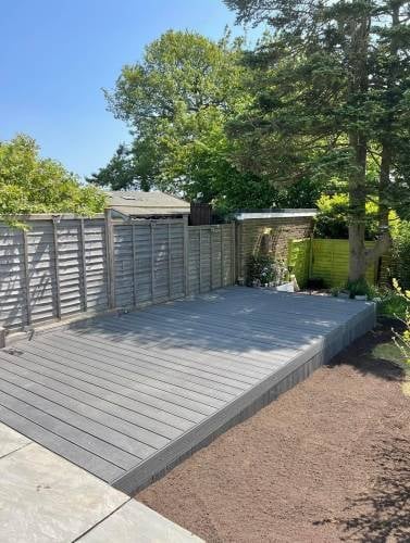 Elevated gray composite deck with slatted fence panels in a backyard garden surrounded by trees on a sunny day