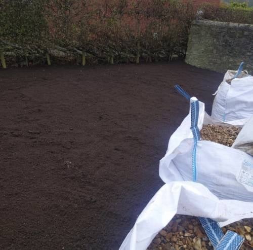White sandbags lined up along dark soil ground with hedges and stone wall in background