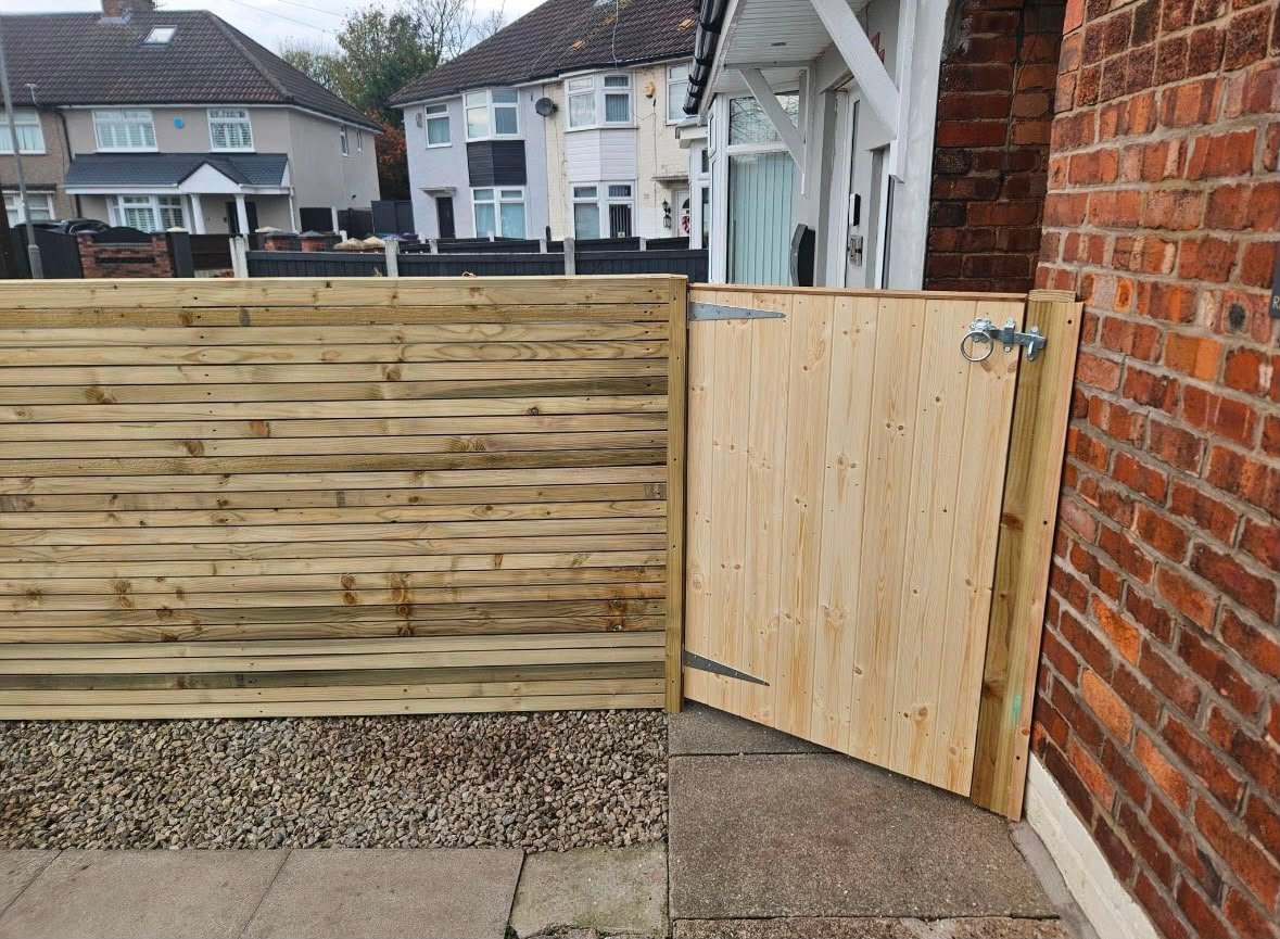 New wooden fence with gate installed in residential driveway next to brick house, with suburban homes visible in background
