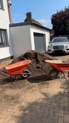 Two red wheelbarrows filled with soil in a residential driveway with a white house and parked car