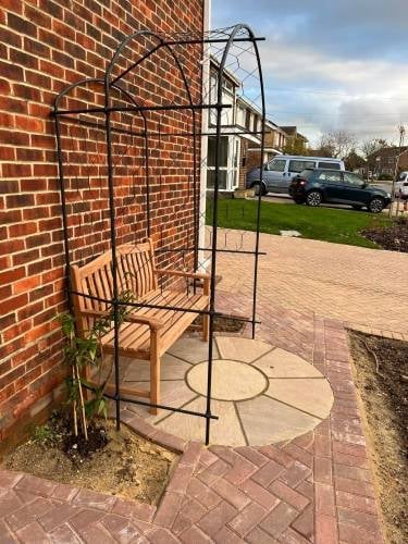 Black metal garden arbor with wooden chair on brick patio next to red brick building