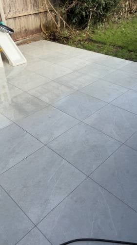 Newly laid gray patio tiles in a residential yard with wooden fence and overgrown vegetation visible in background